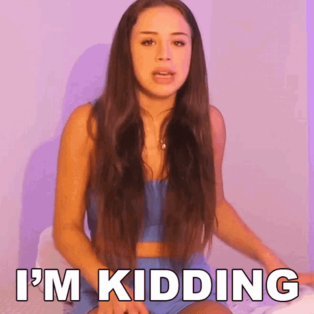a woman with long hair is sitting on a bed and saying i 'm kidding .