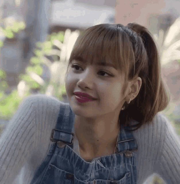a woman wearing overalls and a white sweater is looking at the camera .