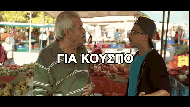 a man in a baseball cap talks to an older man in front of a fruit stand with the words gia koyspo on the bottom