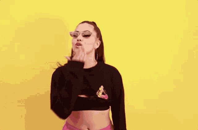 a woman wearing sunglasses and a crop top is blowing a kiss on a yellow background .