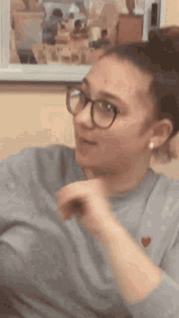 a woman wearing glasses and a gray sweater is sitting in front of a picture on the wall .