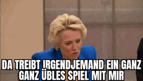 a woman in a blue jacket is sitting in front of a wall and talking in german .