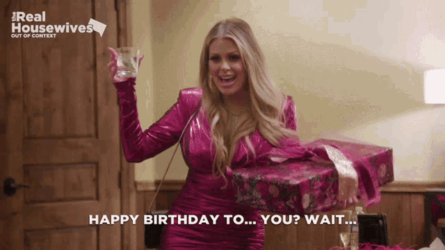 a woman in a pink dress is holding a glass and a gift box and says " happy birthday to you wait "