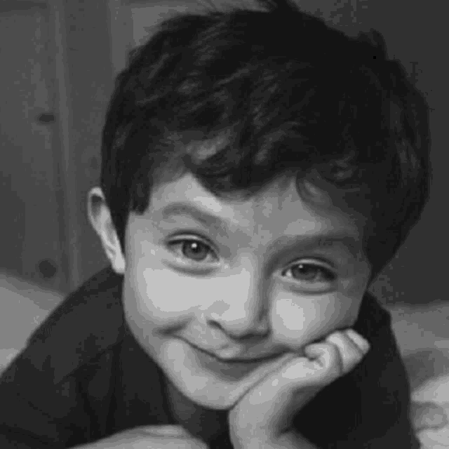 a black and white photo of a young boy smiling with his hand on his face .