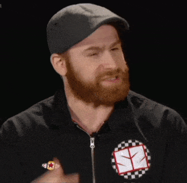 a man with a beard wearing a hat and a black shirt with a checkered y on it