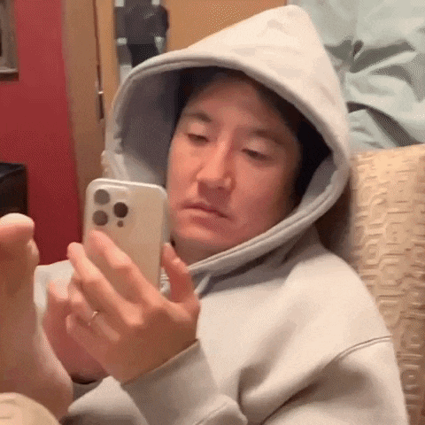 a man in a hoodie is sitting on a couch looking at his cell phone .