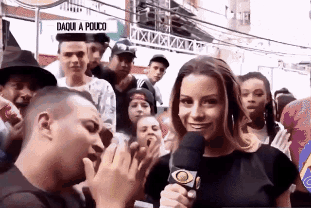 a woman is holding a microphone in front of a crowd and a sign that says daqui a pouco