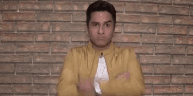 a man in a yellow jacket is standing in front of a brick wall with his arms crossed and looking at the camera .