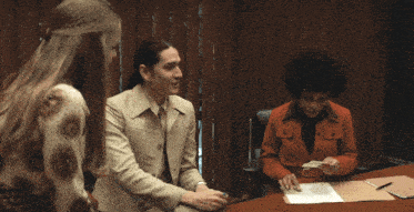 a woman in a red jacket sits at a desk talking to two other women