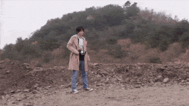 a man in a trench coat is holding a camera in a dirt field