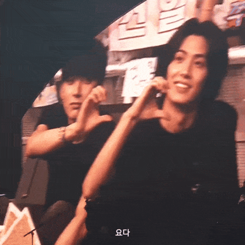 two men are making a heart shape with their hands and one of them is wearing a black shirt with korean writing on it