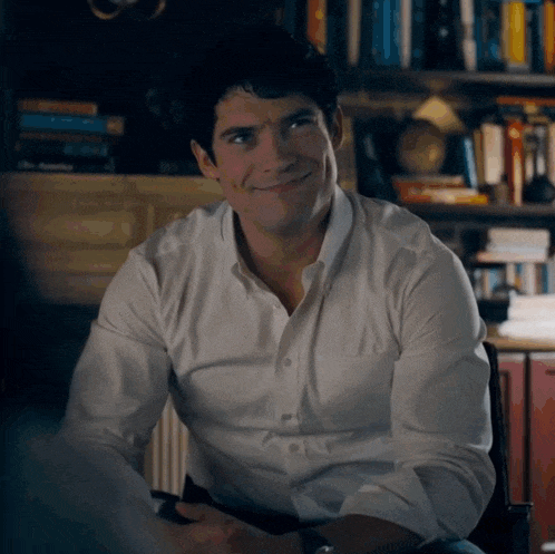 a man in a white shirt is sitting in front of a bookshelf and smiling