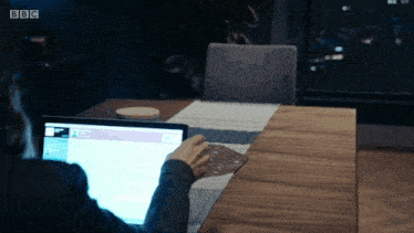 a person sitting at a table using a laptop with the bbc logo on the screen