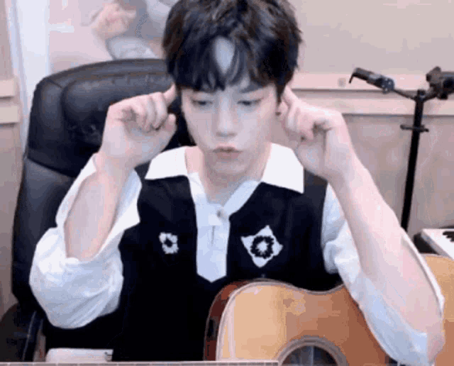 a young man is sitting at a desk with a guitar and covering his ears with his hands .