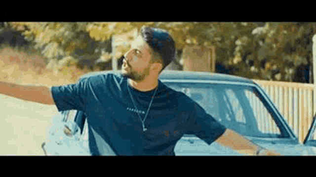 a man in a black t-shirt is standing next to a car with his arms outstretched .