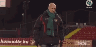 two men are standing next to each other on a soccer field . one of the men is wearing a green scarf .