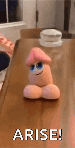 a stuffed penis with a face is sitting on a wooden table and says arise !