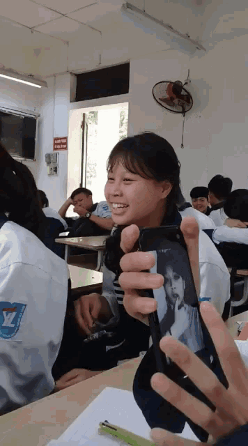 a girl in a classroom holds up a cell phone with a picture of herself on it