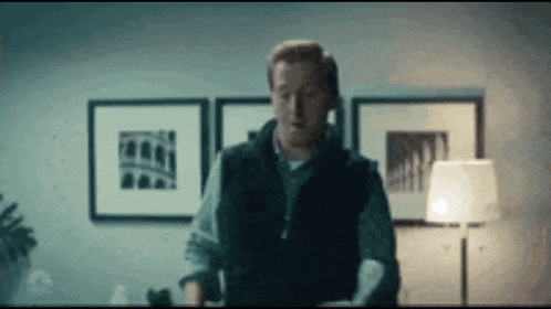 a man in a vest is standing in a room with a lamp