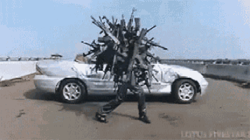 a man is carrying a bunch of guns on his head in front of a silver car .