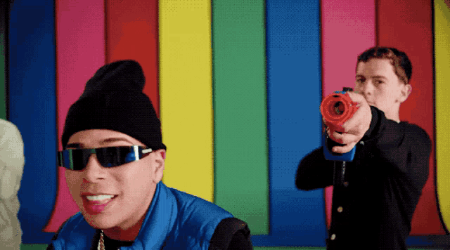 a man wearing sunglasses and a black beanie is holding a toy gun