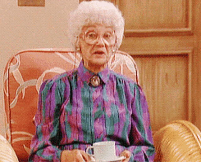 an elderly woman wearing glasses and a purple shirt is sitting in a chair holding a cup of coffee