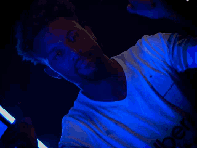 a man in a white t-shirt is standing in a dark room with a blue light behind him