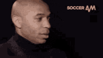 a bald man in a black suit and turtleneck is smiling in front of a black background .