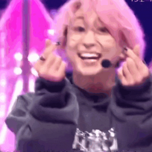 a young man with pink hair is making a heart shape with his hands while wearing a microphone .