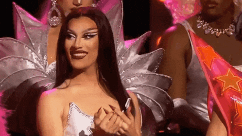 a group of drag queens are standing on a stage and one of them is smiling .