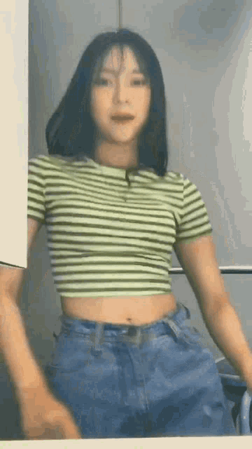 a woman wearing a green striped crop top and blue jeans is standing in front of a mirror .