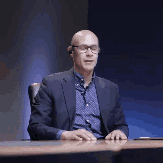 a man in a suit and glasses sits at a table with his mouth open