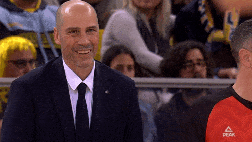 a man in a suit and tie smiles in front of a crowd wearing a peak shirt