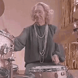 an elderly woman is playing a drum set while wearing a pearl necklace .
