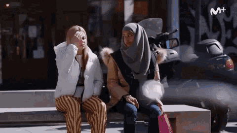 two women sit on a bench with a motorcycle in the background and the letters m + on the bottom
