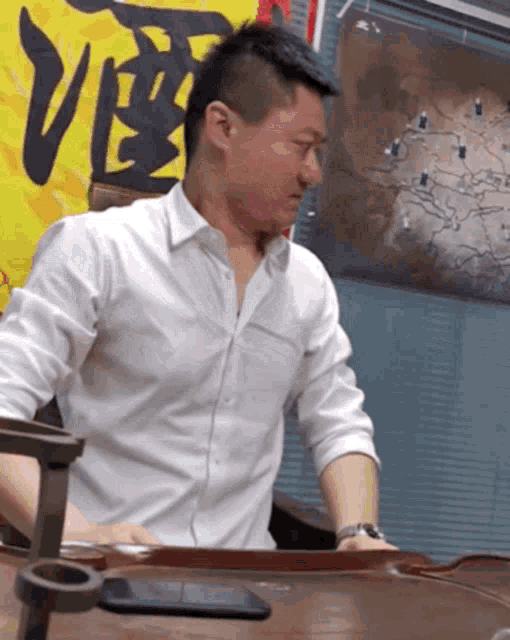 a man in a white shirt sits at a table in front of a wall with chinese writing on it