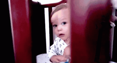 a baby is sitting in a crib and looking through the bars at the camera .