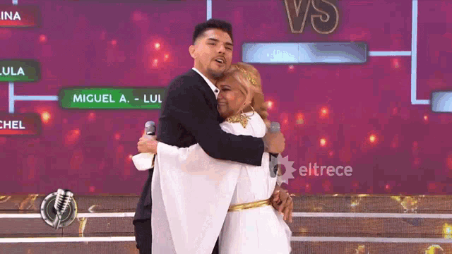 a man in a suit is hugging a woman in a white dress in front of a sign that says eltrece