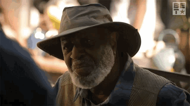 an older man wearing a hat and vest looks at the camera with a be tv logo behind him