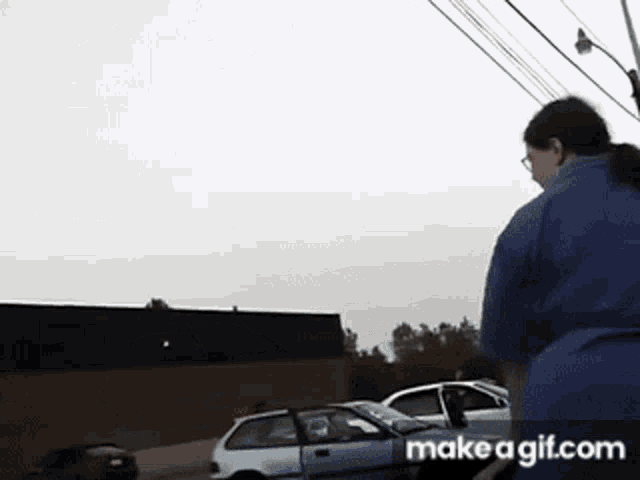 a woman in a blue shirt is standing in front of a white car with make a gif.com written on the bottom right