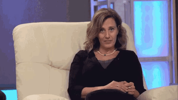 a woman in a black shirt and pearls sits in a white chair