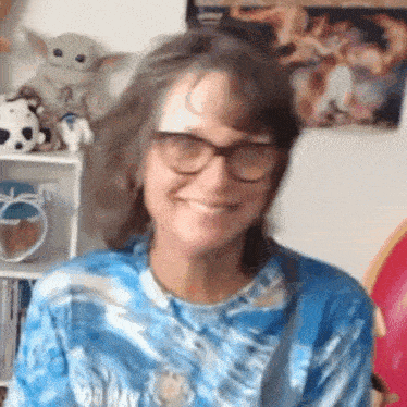 a woman wearing glasses and a blue tie dye shirt is smiling and looking at the camera .