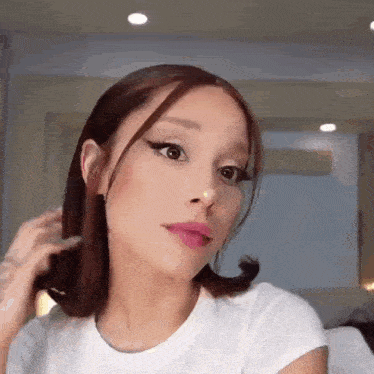 ariana grande is wearing a white t-shirt and holding her hair while looking at the camera .
