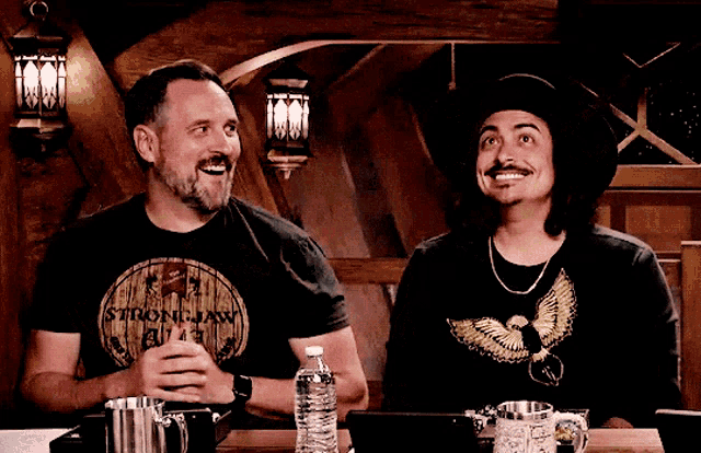 two men are sitting at a table with one wearing a strong jaw shirt