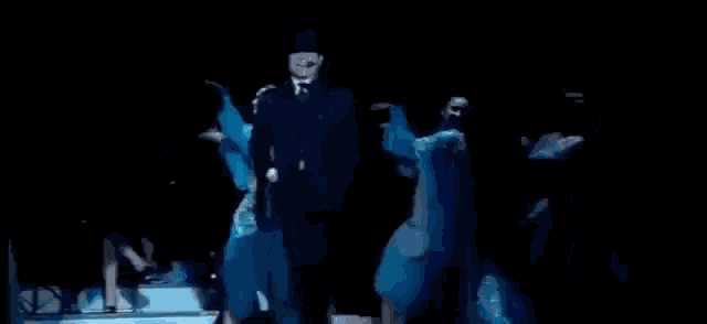 a man in a suit stands in front of a group of dancers with pigliate ' na pastiglia written in white letters