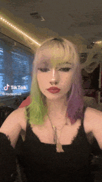a woman with purple and green hair is wearing a black tank top and necklace .