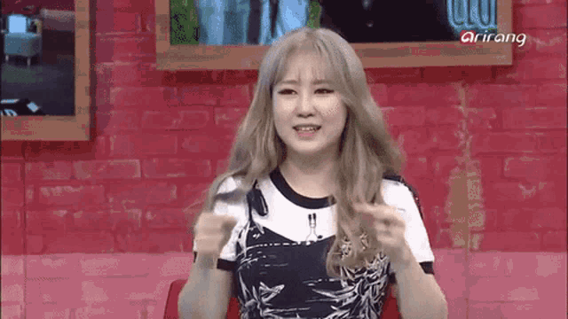 a woman with blonde hair is sitting in front of a red brick wall with a sign that says arirang
