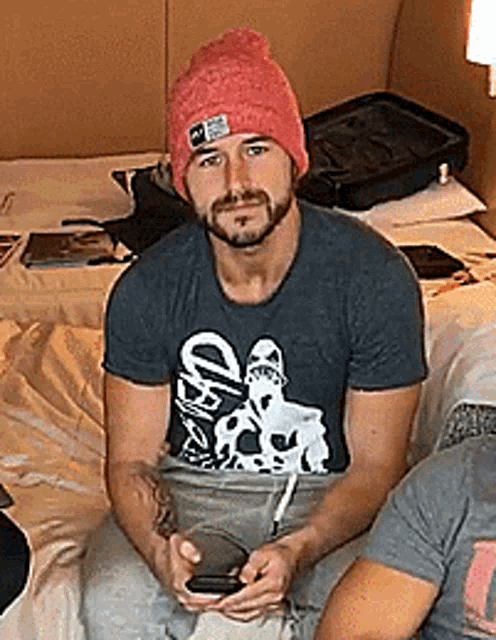a man wearing a beanie and a t-shirt is sitting on a bed holding a cell phone