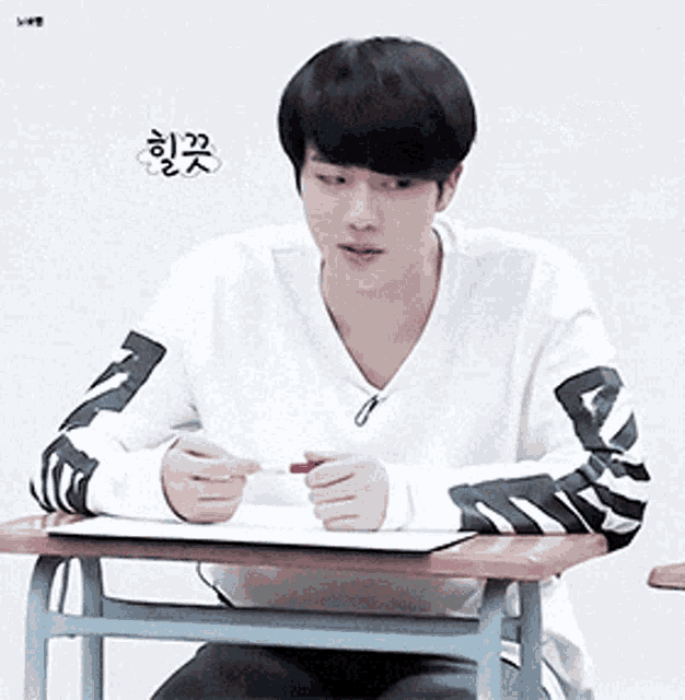 a young man in a white sweater sits at a desk with a pen in his hand