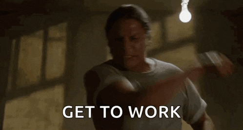 a man is standing in a dark room with a light bulb hanging from the ceiling and says `` get to work '' .